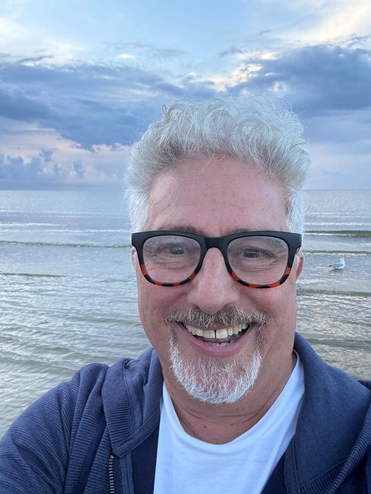 Jerry Fishman smiling at the water's edge, with curly gray hair, glasses and a navy jacket, under a cloudy sky.