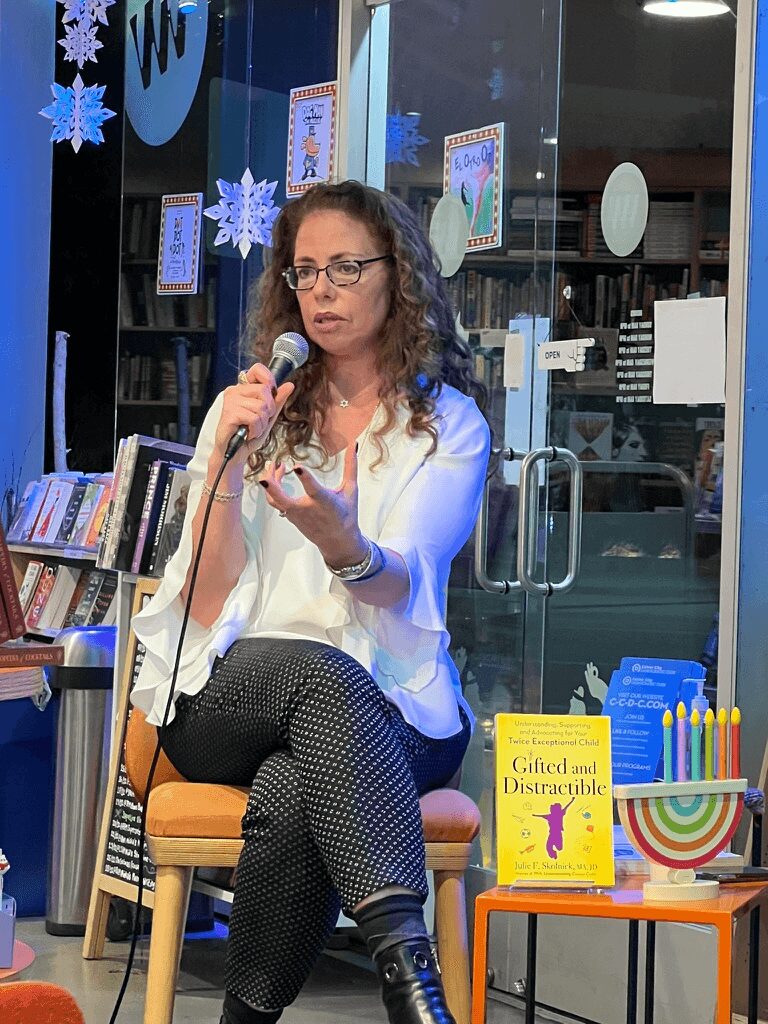 Julie Skolnick speaking into a microphone at a bookstore event, seated beside a copy of her book Gifted and Distractible.